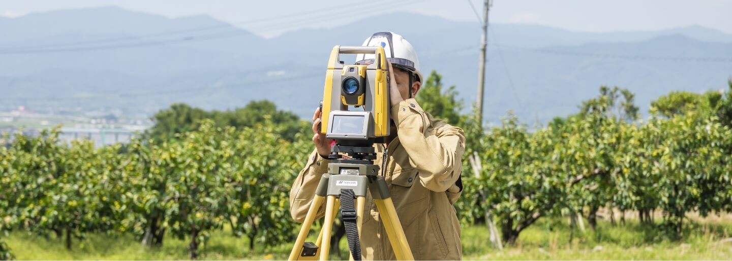 果樹園を背景に測量機器を操作する作業員